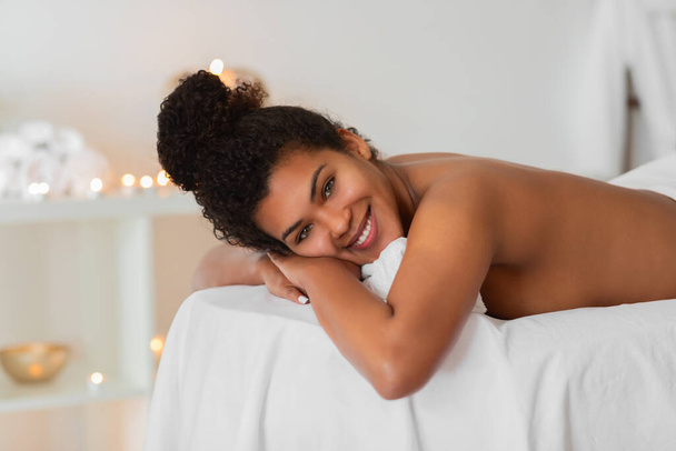 African American γυναίκα βρίσκεται στο στομάχι της σε ένα spa με ένα χαλαρό και ικανοποιημένο χαμόγελο, κοιτάζοντας απευθείας στην κάμερα - Φωτογραφία, εικόνα