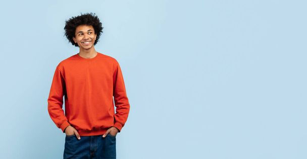 Relaxed african american guy with a subtle smile, comfortably dressed in orange sweater and jeans, standing with hands in pockets against a blue isolated background, copy space - Photo, image