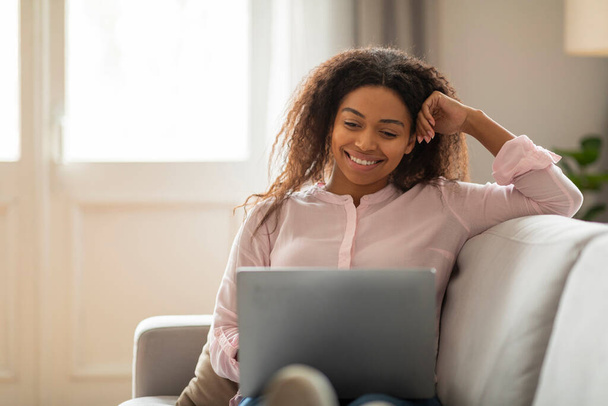 Joven mujer afroamericana alegre usando un ordenador portátil en un sofá cómodo, encarnando la vida laboral flexible en casa - Foto, Imagen
