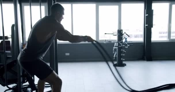 Een atletische man die krachtig traint met zware strijdtouwen in een goed uitgeruste fitnessruimte, die kracht en uithoudingsvermogen laat zien. Atletische man in Gym doet zware touw workout intensief - Video