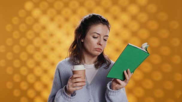 Mujer leyendo libro, pasando página y sosteniendo taza de café desechable, aislado sobre fondo de estudio. Gusano de biblioteca disfrutando de la novela y la bebida con cafeína temprano en la mañana, la cámara B - Imágenes, Vídeo