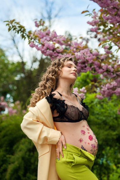 chica joven y rizada embarazada cerca de los árboles flores de cerezo, vientre con adorno de flores, soñadora, en un sentido de paz, elegante traje brillante - Foto, imagen