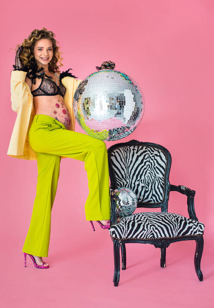 игривая беременная женщина с вьющимися волосами, стоящая у стула. Живот с цветочным узором, в яркой пинап-одежде и нижнем белье, модная студийная фотография на розовом фоне - Фото, изображение
