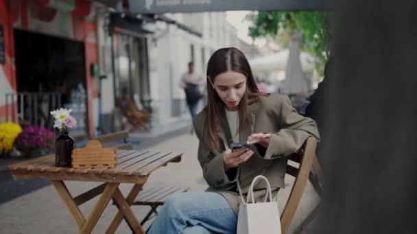 Une jeune femme concentrée vérifie son smartphone alors qu'elle est assise seule à une table de café dans une rue de la ville, incarnant le mode de vie urbain et la connectivité. Jeune femme utilisant un smartphone à la table basse extérieure en ville - Séquence, vidéo