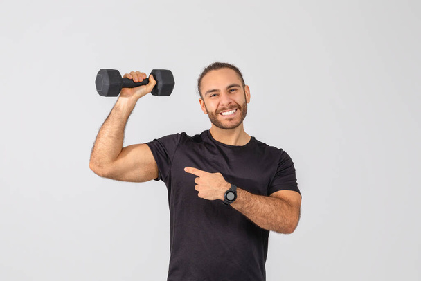 Um homem está de pé enquanto segura um haltere na mão, apontando para o bíceps, mostrando sucesso em sua rotina de exercícios. O homem parece confiante e determinado em seu exercício. - Foto, Imagem