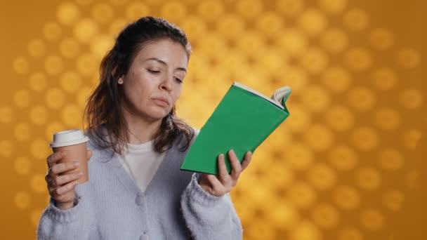Tired woman reading book, holding cup of coffee, isolated over studio background. Exhausted bookworm enjoying novel and caffeine beverage, lacking energy, struggling to stay awake, camera B - Footage, Video
