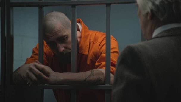 Häftling in orangefarbener Uniform lehnt an Gittern in Gefängniszellen, spricht mit Anwalt und liest Anwaltsvertrag. Häftling verbüßt Haftstrafe für Verbrechen in Justizvollzugsanstalt Gangster in Haftanstalt. - Foto, Bild