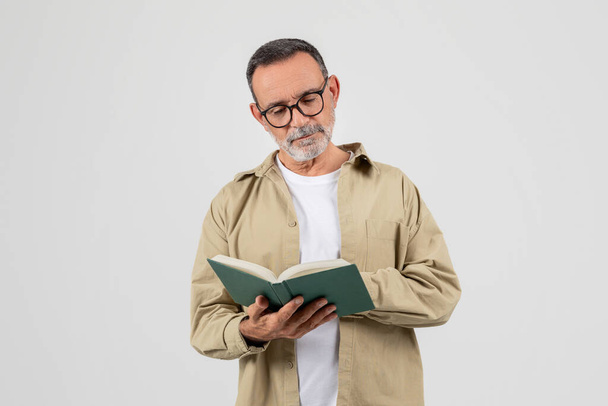 Um homem sênior com barba cheia e óculos está absorto na leitura de um livro. Ele aparece focado enquanto vira as páginas, sentado em uma cadeira dentro de casa. - Foto, Imagem