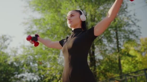 A focused young woman in a black outfit stretches her arms upwards, holding red dumbbells in a sunny park setting. Headphones add to her workout vibe. Woman in Black Workout Gear Stretching Outdoors - Footage, Video