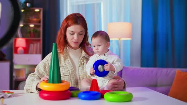 desarrollo del bebé, mujer cariñosa con un niño pequeño recoge una pirámide de plástico de círculos de colores en una habitación acogedora - Metraje, vídeo