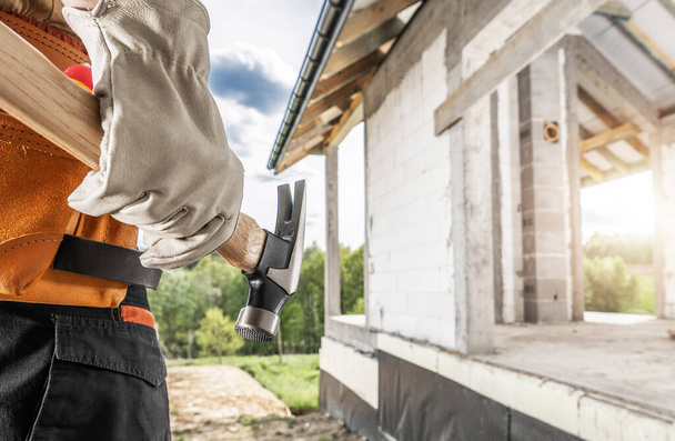 Εργάτης οικοδομών με ένα εργαλείο σφυριών στο χέρι του και το πρόσφατα αναπτυγμένο σπίτι σε ένα υπόβαθρο - Φωτογραφία, εικόνα