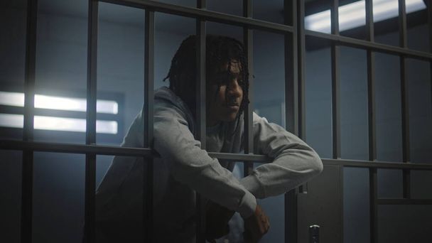 Upset African American teenager with face tattoos stands in prison cell in jail or youth detention center leaning on metal bars. Prison officer passes by young criminal or prisoner in the foreground. - Photo, Image