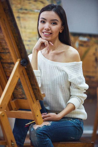 Νεαρή όμορφη γυναίκα καλλιτέχνη ζωγραφικής ενώ εργαζόταν σε ένα στούντιο, χαμογελά στη φωτογραφική μηχανή - Φωτογραφία, εικόνα