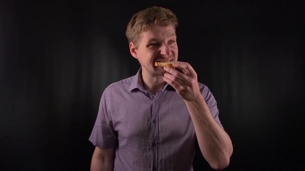 Captivating portrait of a stylish young man with stubble, donning a vibrant purple shirt on a sleek black backdrop, takes bites of meal, exuding an effortless cool vibe. Lifestyle, fashion or culinary - Footage, Video