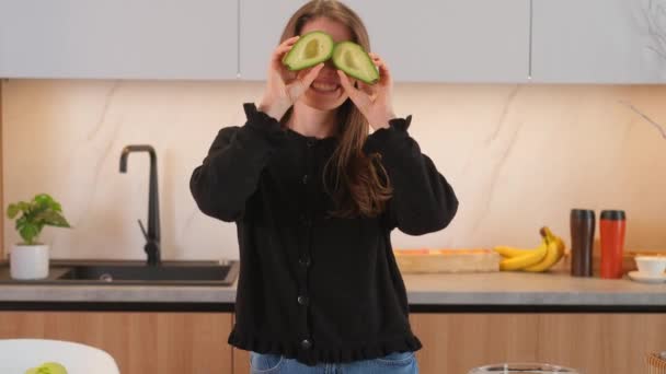 Молода біла жінка тримає нарізане авокадо на кухонному фоні. - Кадри, відео