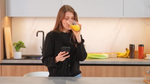 Bájos fiatal nő sms-ező okostelefon és ivás pohár narancslé a konyhában. - Felvétel, videó