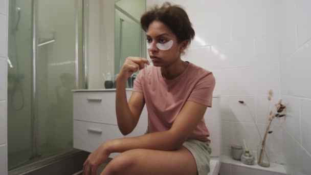 Młoda chora dziewczyna z przepaskami pod oczami siedzi na toalecie w łazience i myje zęby rano - Materiał filmowy, wideo