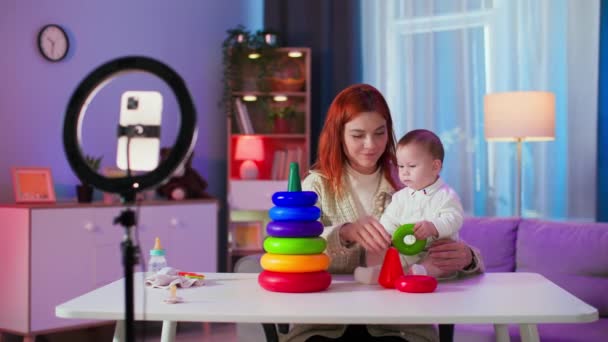 babyblog, söpö naisbloggaaja, jolla on pieni lapsi, tallentaa videon älypuhelimella sosiaalisille verkostoille istuessaan viihtyisässä huoneessa - Materiaali, video