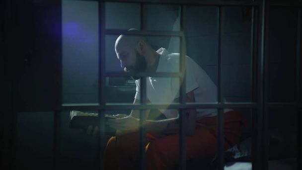 Männlicher Häftling in orangefarbener Uniform sitzt auf dem Bett in der Gefängniszelle, liest die Bibel. Illegal verurteilter Mann verbüßt Haftstrafe im Gefängnis Blick durch Metallstangen. Haftanstalt oder Justizvollzugsanstalt. - Foto, Bild