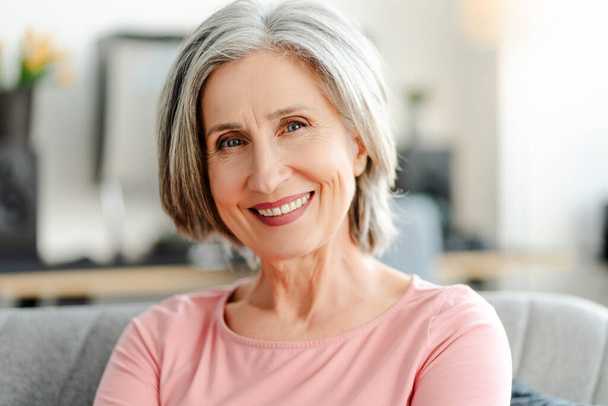 Glückliche, selbstbewusste Seniorin, die auf dem bequemen Sofa zu Hause in die Kamera blickt. Porträt lächelnde Rentnerin mit eleganten grauen Haaren, weißen Zähnen. Natürliche Schönheit, gesundes Lebensstil-Konzept - Foto, Bild