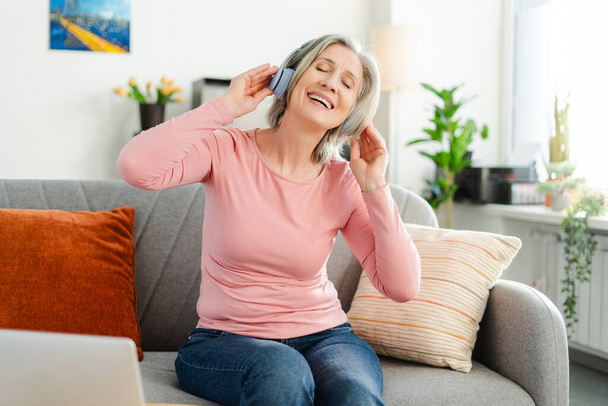 Attractive happy senior woman listening music in wireless headphones, having fun sitting on comfortable sofa at home. Technology, positive lifestyle concept  - Photo, Image