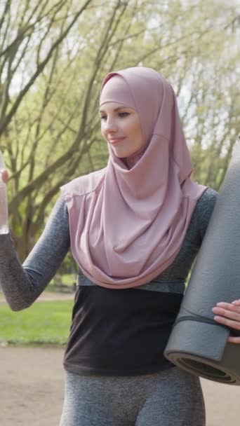 Schermo verticale: Donna sana e sportiva in hijab che tiene il tappetino di yoga e l'acqua della bottiglia mentre si trova al parco cittadino. Sorridente musulmana femminile in pausa durante l'allenamento all'aria aperta. - Filmati, video