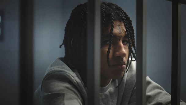 Yüzünde dövmeler olan kızgın Afrikalı Amerikalı genç mahkum hapisteki hücrede duruyor ve kameraya bakıyor. Genç suçlu, suçtan hapis cezasına çarptırıldı. Islahevi. Portre. - Fotoğraf, Görsel