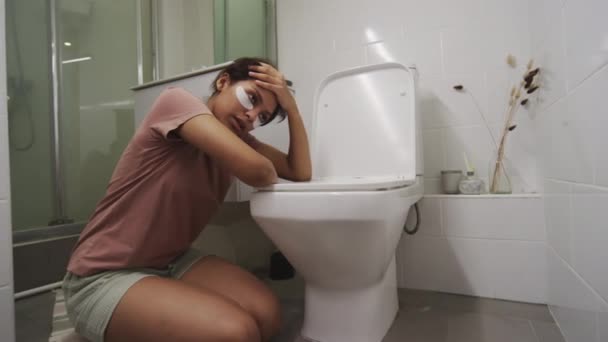 Sairas teini-ikäinen tyttö, jolla on silmälaput, istuu lattialla kylpyhuoneessa, nojaa wc: hen ja hengittää voimakkaasti, kun kärsit ruokamyrkytyksestä - Materiaali, video