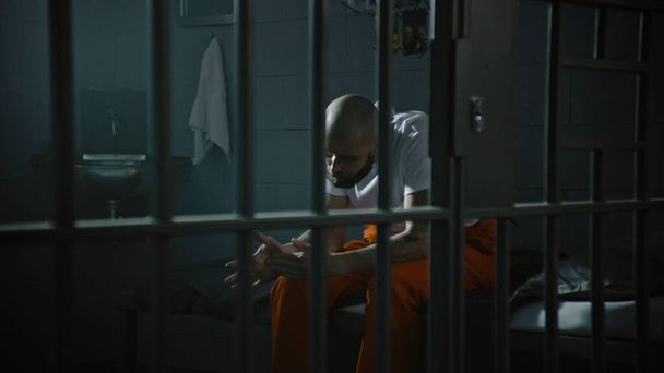 Crimineel in oranje uniform zit op het celbed. Gevangene zit gevangenisstraf uit voor misdaad in de gevangenis. Gangster in detentiecentrum, gevangenis. Justitie. Zicht door metalen staven. - Foto, afbeelding