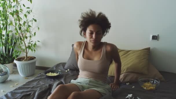 Młoda dziewczyna z brudne włosy budzi się na łóżku nieczyste łóżko z rozrzuconych przekąsek i czuje się źle rano po imprezie w domu - Materiał filmowy, wideo