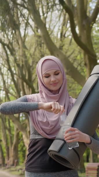Vertikální obrazovka: Atraktivní muslimka ve sportovním oblečení pije sladkou vodu z láhve po ranním cvičení v zeleném parku. Hezká fena v hidžábu drží v rukou podložku na jógu. - Záběry, video