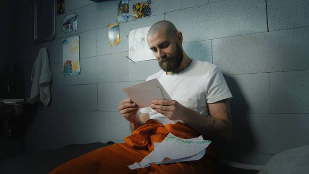 Overstuur mannelijke gevangene kijkt naar familiefoto 's en kindertekeningen zittend op bed in de cel. Crimineel zit gevangenisstraf uit voor misdaad in de gevangenis. Een detentiecentrum of een gevangenis. Portret. - Foto, afbeelding