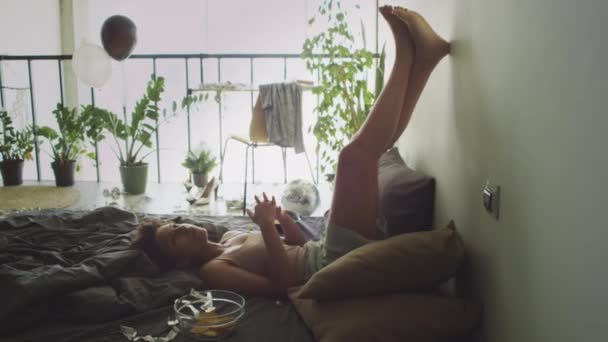 Ragazza esausta sdraiata su un letto disordinato con le gambe alzate sul muro e sentirsi male durante il giorno a casa - Filmati, video