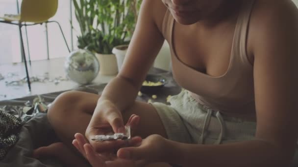 Plan incliné de la fille prenant des pilules tout en étant assis sur un lit désordonné non fait et se sentant malade le matin à la maison - Séquence, vidéo