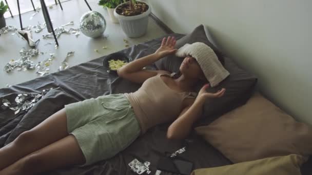 Hoge hoek uitzicht van ziek meisje lijdt aan hoofdpijn zetten natte handdoek op het voorhoofd, terwijl rusten op onopgemaakt bed in rommelige kamer met verspreide snacks, discobal en knutselen - Video