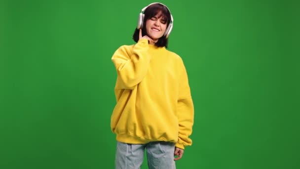 Young woman in modern oversize outfit listening music in headphones and dancing against green studio background. Concept of human emotions, fashion and beauty, style, technology. ad - Footage, Video