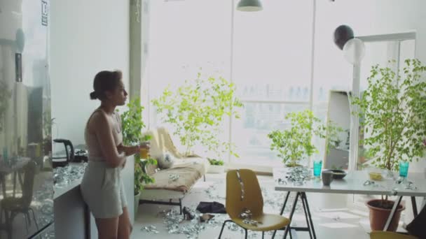 Młoda zmęczona dziewczyna chodząca w brudnym salonie z świecidełkiem na podłodze, pijąca sok i sprawdzająca wiadomości przez telefon komórkowy rano w domu - Materiał filmowy, wideo