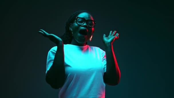 Překvapená, vzrušená mladá Afroameričanka se drží za ruce na tvářích emocí v neonovém světle na pozadí studia. Pojem lidské emoce, krása a móda, styl, - Záběry, video
