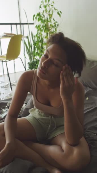 Portrait vertical de jeune fille somnolente assise sur un lit non aménagé dans une pièce encombrée, frottant son visage et regardant la caméra - Séquence, vidéo