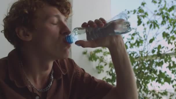 Junger hungriger Mann trinkt Wasser, SMS auf Handy und lehnt sich auf Couch zurück, während er sich nach der Party zu Hause schlecht fühlt - Filmmaterial, Video