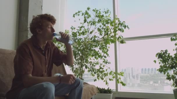 Nuori gen Z mies kärsii krapula juomaveden ja tarkistaa puhelimen istuessaan sohvalla sotkuinen huone jälkeen kotibileet - Materiaali, video