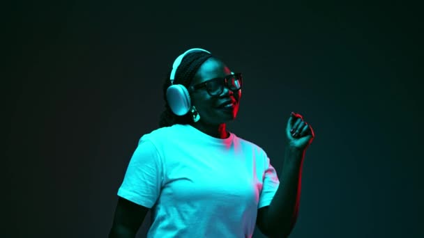 Donna afro-americana rilassata che ascolta musica in cuffia e canta in luce rossa al neon sullo sfondo dello studio di gradiente. Concetto di emozioni umane, bellezza e moda, stile, - Filmati, video