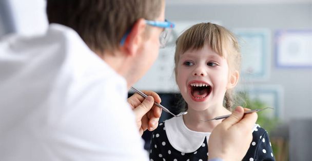 Male dentist look at open mouth litle happy girl child againlt hospital office background portrait. Tooth treatment concept - Photo, Image
