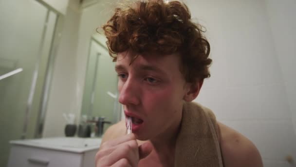 Нахилений знімок виснаженого молодого чоловіка з кучерявим волоссям, що сидить на туалеті, чистить зуби і протирає обличчя рушником, маючи погане похмілля вранці вдома - Кадри, відео