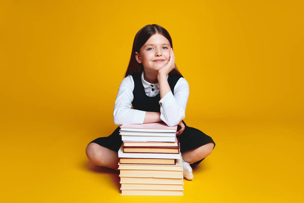 Vrolijk basisschoolmeisje in uniform, zitten in de buurt bos van boeken met gekruiste benen en de hand onder de kin, geïsoleerd op gele achtergrond - Foto, afbeelding