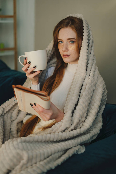 Hermosa joven pelirroja temprano en la mañana, leyendo un libro, disfrutando del café caliente de la taza, sentada en un cómodo sofá. Ambiente doméstico acogedor - Foto, Imagen