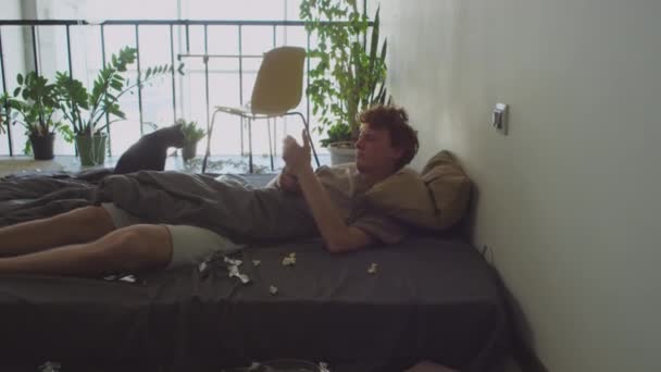 Jeune homme affamé se réveillant dans un lit sale, vérifiant le téléphone mobile, bâillant et buvant de l'eau de la bouteille le matin après la fête à la maison - Séquence, vidéo