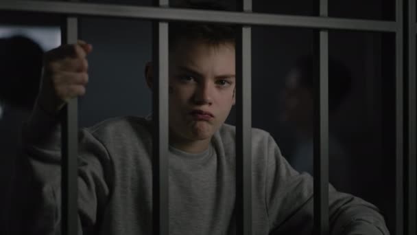 Kavkazský teenager s tetováním na obličeji stojí ve vězeňské cele ve vězení, drží kovové mříže a dívá se do kamery. Různí mladí vězni mluví v pozadí. Středisko pro mladistvé nebo nápravné zařízení - Záběry, video