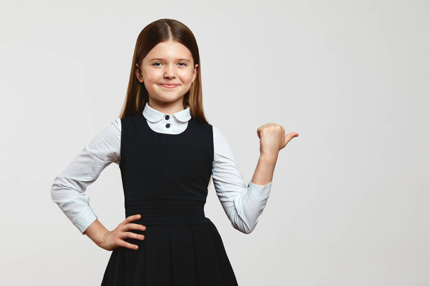 Cute kid girl in school uniform looking at camera, keeping hand on waist and pointing at free empty space for text, isolated against white background - Photo, Image