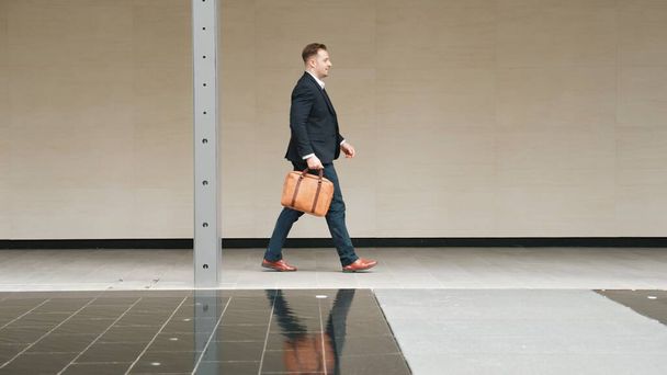 Πλευρική άποψη των επιχειρηματιών κρατώντας βαλίτσα και περπατώντας στο χώρο εργασίας κατά μήκος του δρόμου στην αστική πόλη. Επαγγελματίας διευθυντής έργου πρόκειται να συναντηθούν, ενώ φοράει επίσημη κοστούμι τα πόδια στο εξωτερικό. Ουρμπάνος. - Φωτογραφία, εικόνα